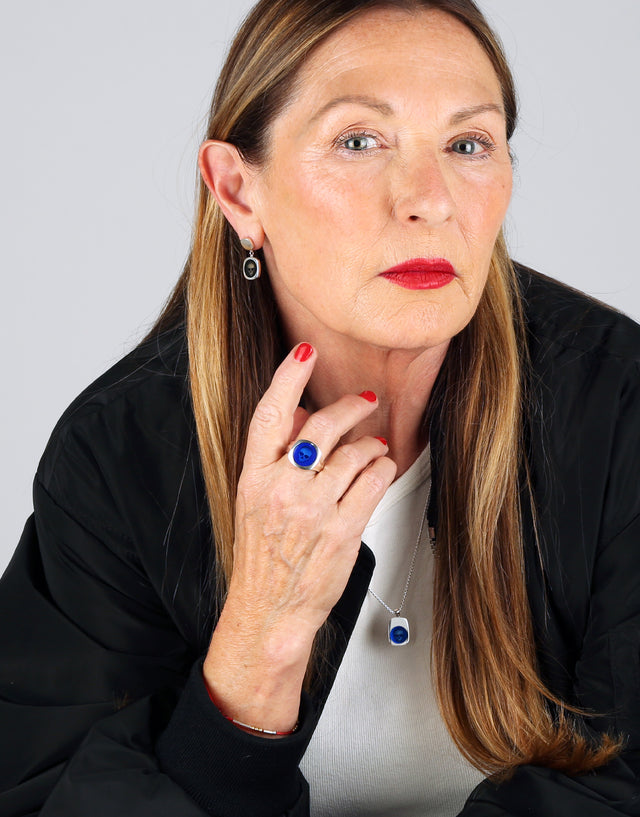 A woman is wearing a signet set of earrings, ring and necklace with black and blue enamel and a skull design on them.